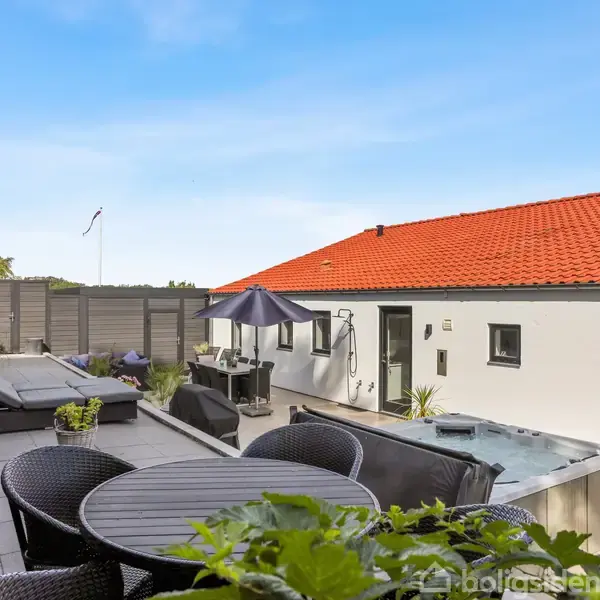 Rundt havebord står på terrasse ved siden af jacuzzi. Solsenge og parasol markerer afslapningsområdet, flankeret af et hvidt hus med rødt tag, omgivet af højt hegn og grønne planter.