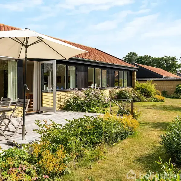 En gul murstensvilla med rødt tegltag og store vinduer. En åben terrassedør fører ud til en have med græs og blomsterbede. En parasol og havemøbler står på terrassen.