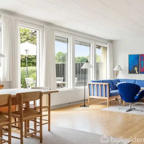 Stue med trægulv og store vinduer, fyldt med naturligt lys. Spisebord med stole og blå sofa ved siden af kunst på væggen. Terrasse udenfor.