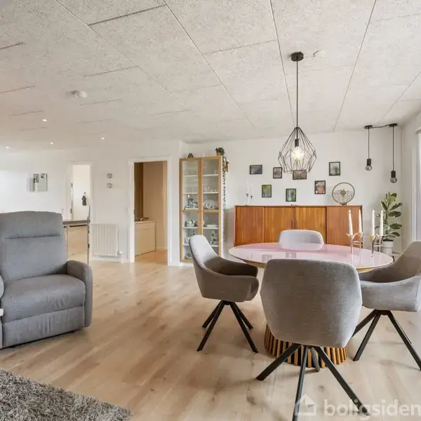 Stue med grå lænestol og spisebord omgivet af fire grå stole. Hængelampe over bordet. Store vinduer til højre giver lys til rummet med planter og dekorative elementer.
