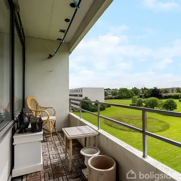 Balkon med træmøbler og planter. Ovenpå et flisegulv står en bord og en stol. Udsigt over en grøn græsplæne og buske under en blå himmel.