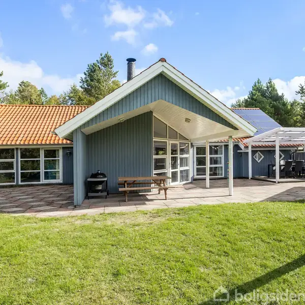 Et blåmalet sommerhus med store vinduer og et rødt tegltag står stille. Det er omgivet af en terrasse og en grøn græsplæne med skov i baggrunden.