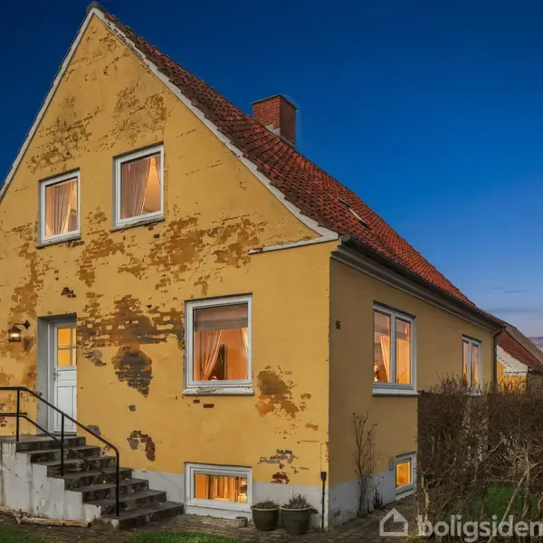 Et gult murstenshus med rødt tegltag, lidt falmet ydermur. Huset har fem oplyste vinduer og en trappe ved hoveddøren. Det står i en have omgivet af buske.