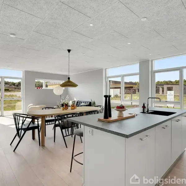 Spisebord med sorte stole står i en moderne stue, oplyst af store vinduer. Køkkenø med barstole står til højre, omgivelserne er lyse og minimalistiske.