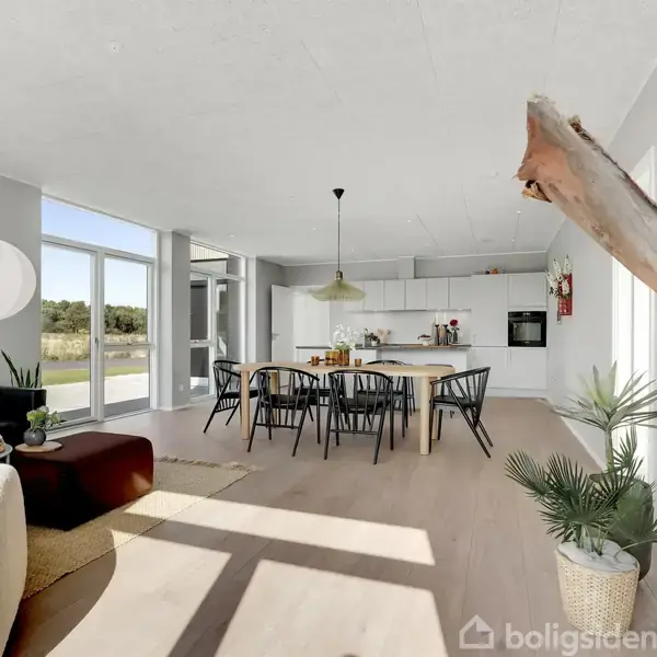 Et moderne spisestue interiør med et lyst træbord omgivet af sorte stole, en gul lampe hængende over, med udsigt til en grøn have gennem store vinduer. Bordet er dekoreret med blomster.