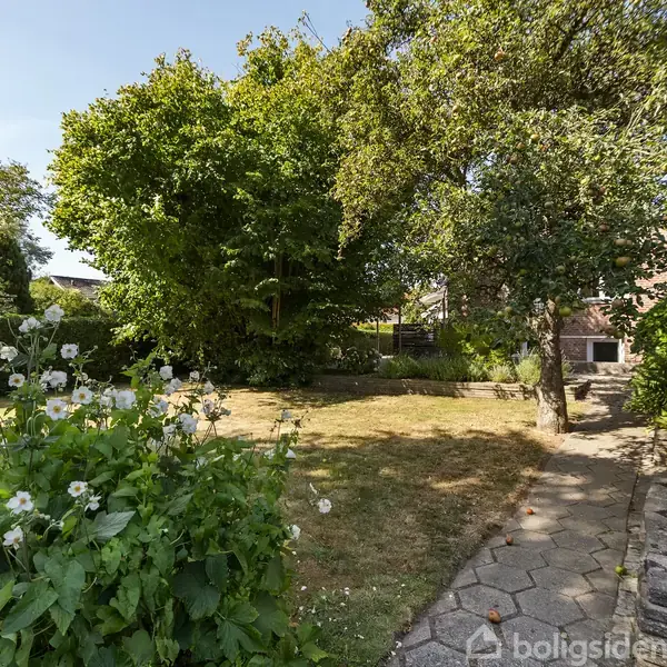 En have omgivet af frodige træer og buske. Foran er der et blomsterbed med hvide blomster. En flisebelagt sti fører gennem græsplænen til et hus.
