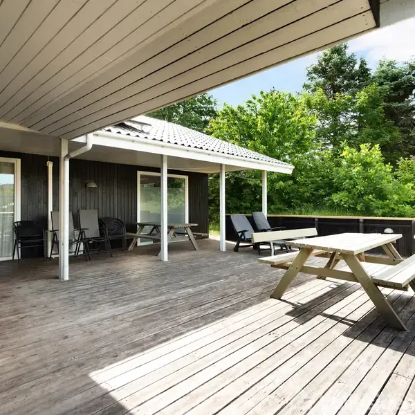 Terrassen har en træbænk og grill, omgivet af grønne træer. Flere loungestole står under et halvtag, og solen skinner på den lyse træoverflade.