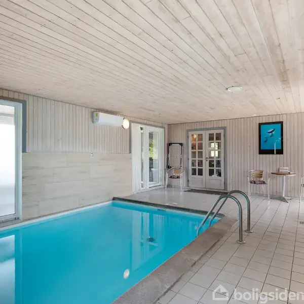 En indendørs swimmingpool fyldt med klart vand, omgivet af en lys træbeklædt væg og loft. Ved siden af poolen er der en lille siddegruppe med stole og et bord.