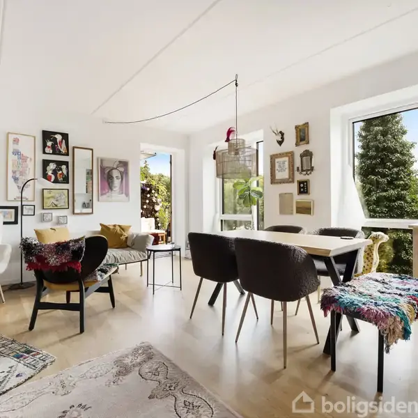 Stue med lyse trægulve fyldt med kunstværker på væggen; sofagruppe til venstre, spisebord med stole til højre, store vinduer med udsigt til grøn have.