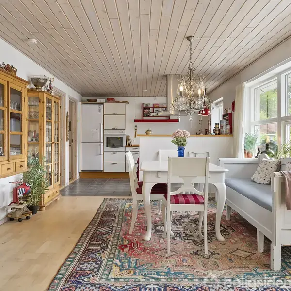 En stue med et hvidt spisebord og stole er opstillet på et farverigt tæppe. Der er en træreol med glaslåger til venstre og store vinduer til højre.