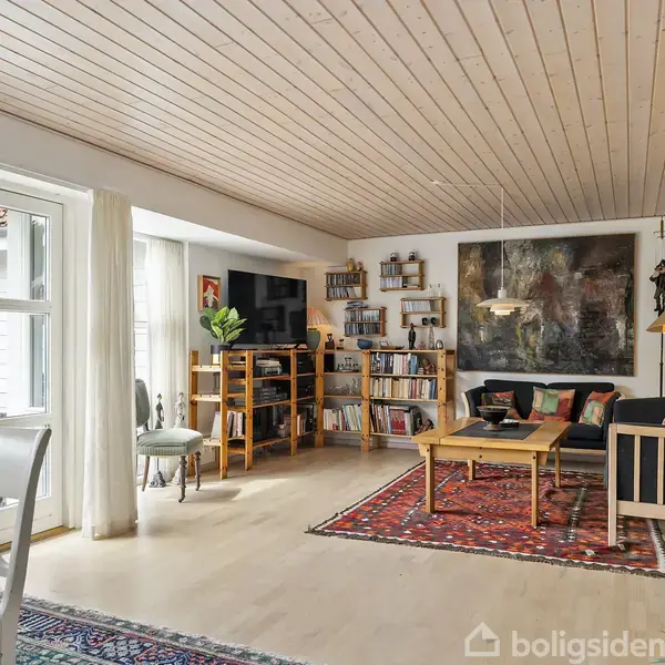 Stue indrettet med sofa og sofabord på et rødt tæppe, flankeret af bogreoler. Store vinduer lader lys ind, træloft tilføjer varme. Maleri hænger over sofaen, tekst "Boligsiden" ignoreret.