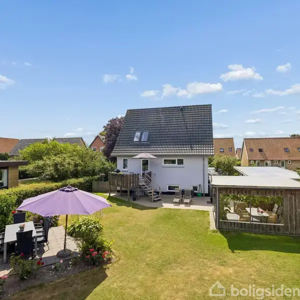 Hus med grå tag og hvide vægge står i en have med græsplæne. Terrasse med parasol og siddepladser til venstre, overdækket siddeområde til højre. Solrig himmel.