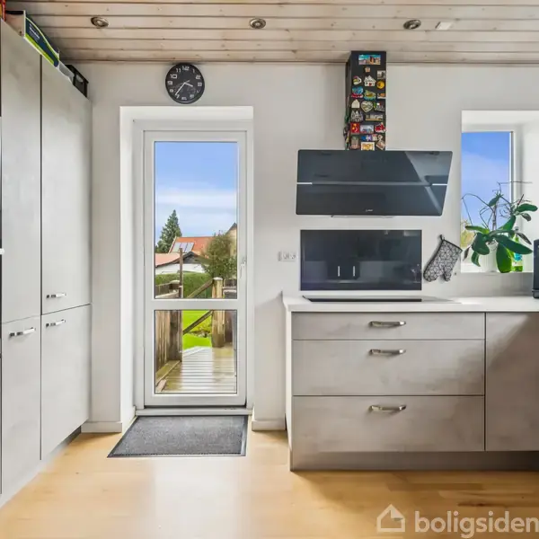 En hvid to-etagers hus med gråt tag har en træterrasse med trapper ned til en grøn have. En træstol og bord står på terrassen. Naboer og biler ses i baggrunden.