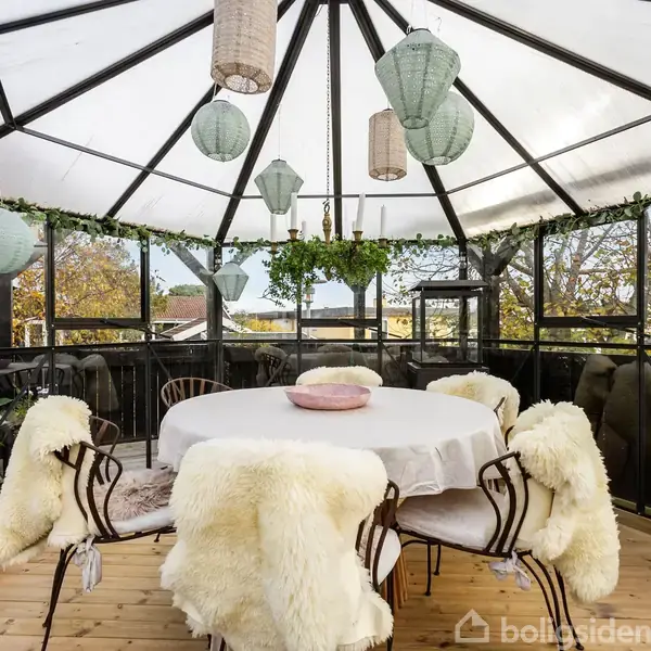 Rundt bord med hvid dug og seks stole med pelsdækket ryglæn står i en glaspavilion. Grønne planter og lanterner hænger fra loftet, omgivet af solrigt landskab udenfor.