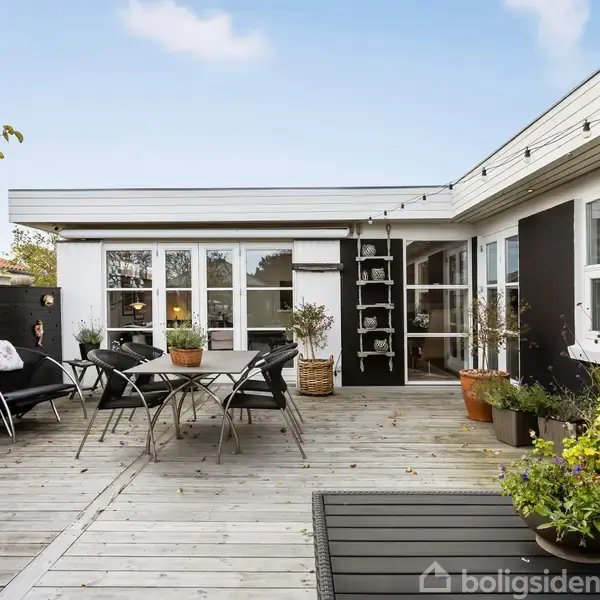 En træterrasse med havemøbler og planter i potter omkranser et moderne hus med store vinduer under blå himmel. Spisebord og stole står centralt på terrassen.