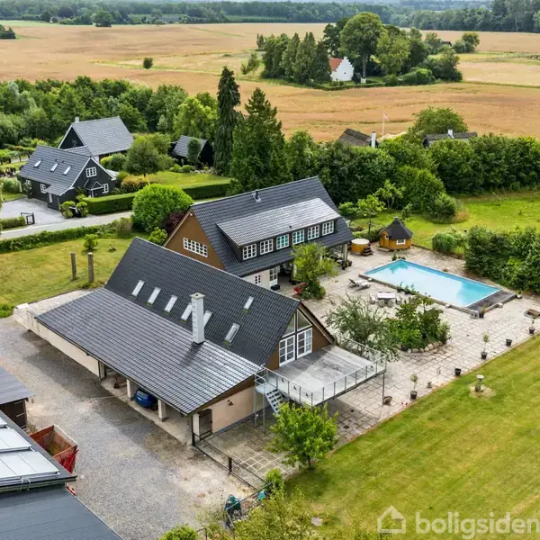 Stor ejendom med to bygninger, en swimmingpool og terrasse, omkranset af græsplæner. Beliggende i landligt område med marker og nogle træer i baggrunden.
