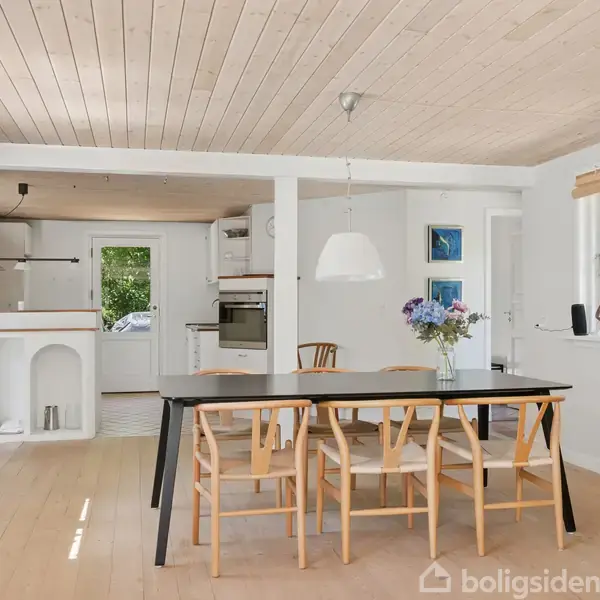 Et spisebord med seks stole står i en lys, moderne spisestue. En skål med blomster pynter bordet. Til venstre ses et køkken med åbne hylder og ovn.