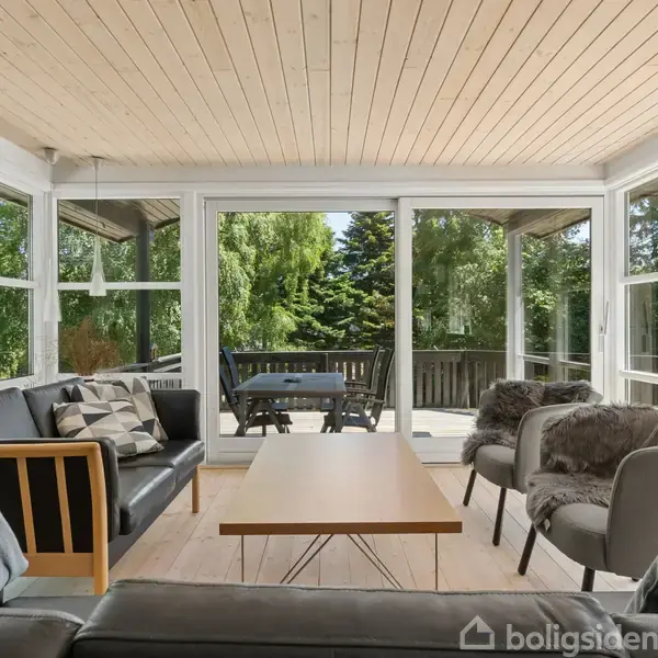 Stue med sofaer og stole omkring et sofabord, handling kan være afslapning eller samtale. Omgivet af store vinduer med udsigt til en terrasse og frodig have.
