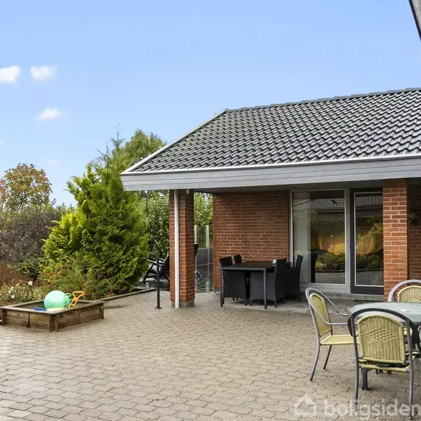 En terrasse med havemøbler står på belægning foran et rødt murstenshus med sort tag. I forgrunden er der en rund bordgruppe. Haven rundt om er frodig og grøn.