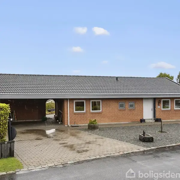 En rødstensbungalow med mørkt tag står stille i en forstadshave omgivet af grus og fliser med en velplejet hæk på venstre side. En autocamper ses i baggrunden.