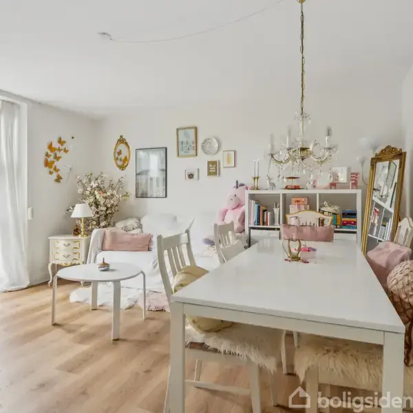 Stue med hvidt spisebord og stole, pyntet med en lysekrone. Ved siden af ses en sofa, sofabord og bogreol. Store vinduer i baggrunden giver naturligt lysindfald.