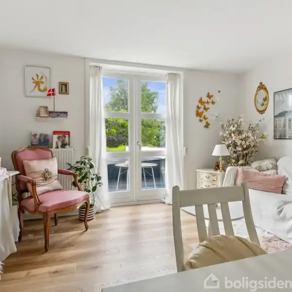 Stue med en hvid sofa, et lille bord, og en stol. Døre fører til en terrasse med haveudsigt. Dekoreret væg med billeder, planter, og lampe.