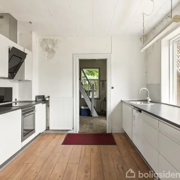 Hvidt køkken med sorte bordplader, komfur med emhætte, og vask. Et stort vindue til højre, trægulv og rød måtte. Døråbning til rum med vindue og synlige træbjælker.