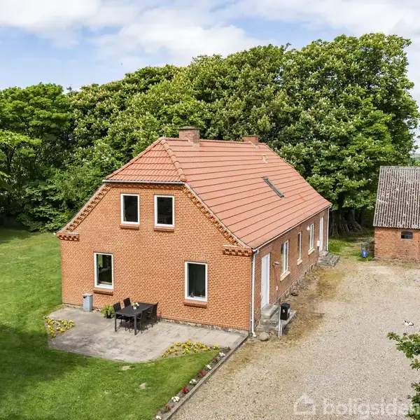 Et rødt murstenshus med rødt tegltag står på en stor grøn græsplæne. Til venstre er et lille terrasseområde med bord og stole. Til højre ses en mindre udbygning. Baggrunden domineres af tæt skov.