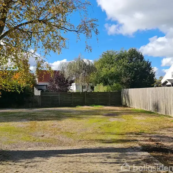 En åben gårdsplads med brosten og græs, omringet af træer og hække, og et hegn løber langs den ene side. En birketræ står i forgrunden.