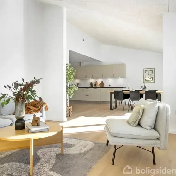 Stue med moderne møbler: to sofaer, sofabord, og dekorative puder. Lys fra vinduer. I baggrunden ses køkken med spisebord og stole. Grøn plante tilføjer liv til rummet.
