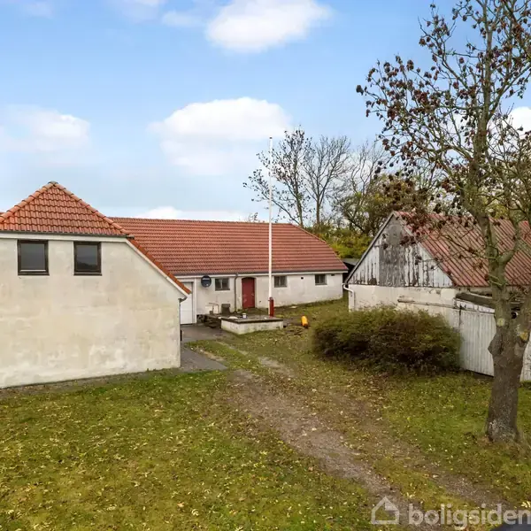 Hus med rødt tegltag på en grøn grund, flankeret af træer og buske. En klart markeret sti fører til hovedindgangen. Skylinen er blå med lette skyer.