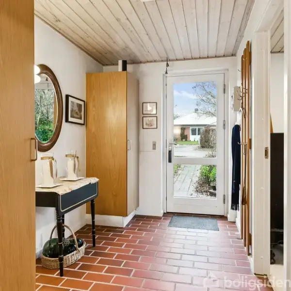 Entredør med glas åbner mod gang med rødlige klinker. Rundt spejl over sort bord står til venstre. Træloft og billeder hænger på væggen.