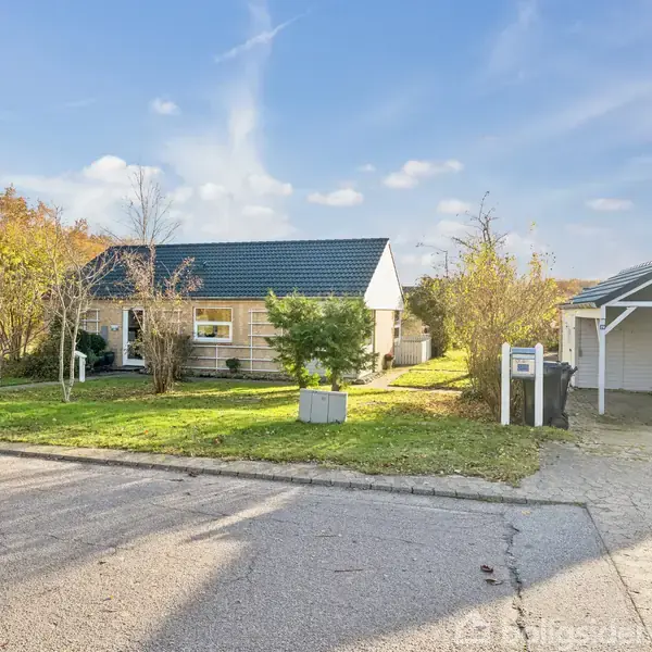 Et hus med mørkt tag står på en grøn græsplæne omgivet af træer og buske. Ved siden af er en carport, og der er en indkørsel foran.