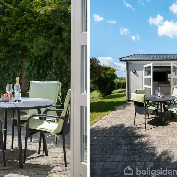 Et rundt havebord med vin og glas står på en flisebelagt terrasse i sollys, omgivet af grønne buske og et hvidt hus med åbne døre.
