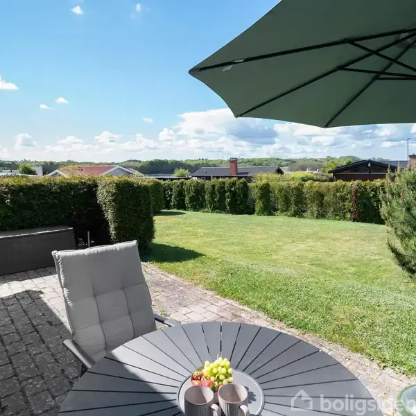 En rund havebord står på en flisebelagt terrasse med en parasol, omgivet af grønne hække. Solskinsdag i et boligområde med huse i baggrunden.