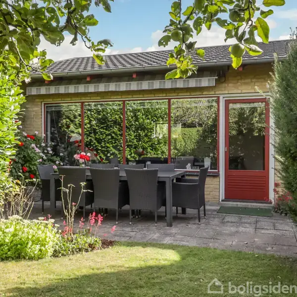 Terrasse med sort spisebord og stole, omgivet af frodig have og grønt buskads. Indgang med rød dør og store vinduer åbner sig mod huset.