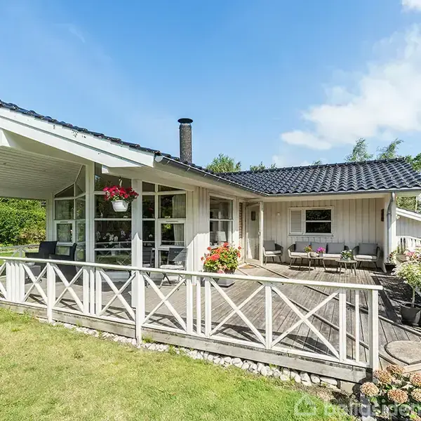 Et hvidt sommerhus med en overdækket terrasse og sort tag står i en grøn have. Blomster hænger fra taget, og havemøbler er placeret på terrassen.