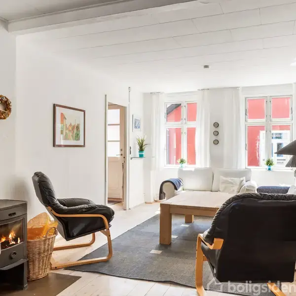 Brændeovn med flammer, står i en stue med to lænestole og et sofabord. Området har lyse vægge, store vinduer med røde gardiner og flere dekorative elementer.