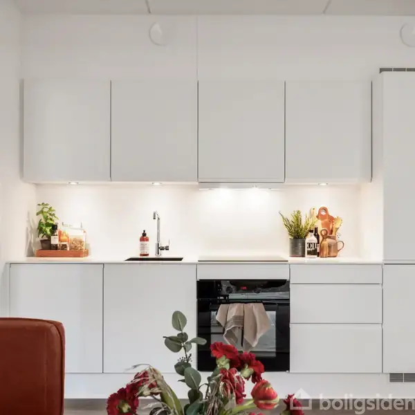 Hvidt køkken med lukkede skabe og indbygget ovn. Plante og køkkenredskaber på bordpladen. Lyse omgivelser med rødbrune blomster i forgrunden.