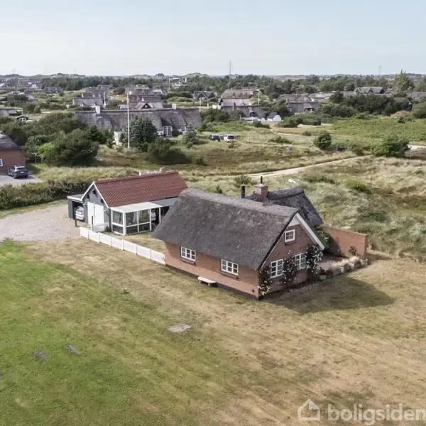 Et stråtækt hus står frit på en åben græsmark. I baggrunden ses flere spredte huse omgivet af grønt landskab og buskads.