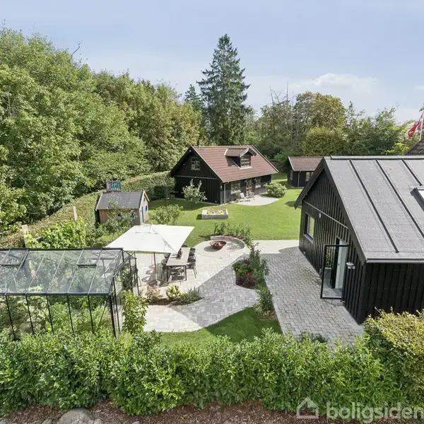 Et idyllisk haveområde med et sortmalet hus, drivhus, og terrasse med parasol og bord. Omgivet af frodige træer og grønne buske giver det en hyggelig atmosfære.