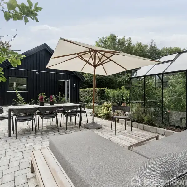 En terrasse med en grå sofa og et bord under en parasol. I nærheden står et drivhus og et sort hus i en haveomgivelse med brostensbelægning og frodige planter.