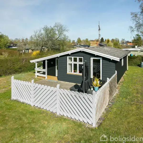 Et lille sort træhus står stille omgivet af en grøn græsplæne og hæk. Foran huset er en hvidmalet træterrasse med blomster og havemøbler.