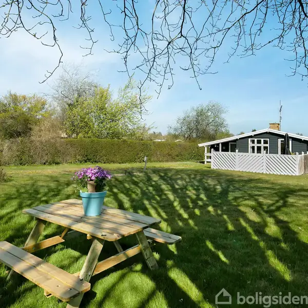 Terrassemøbler står på en flisebelagt gårdhave. Et bord med stole omkring er ved siden af en grill og en parasol. Omkranset af et hvidt plankeværk og et træ står til venstre.