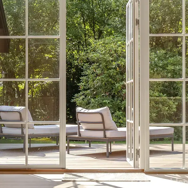 To liggestole står stille på en terrasse, omgivet af grønne træer. Store glasdøre åbner op til rummet, hvilket giver et klart kig til den solrige udendørs have.
