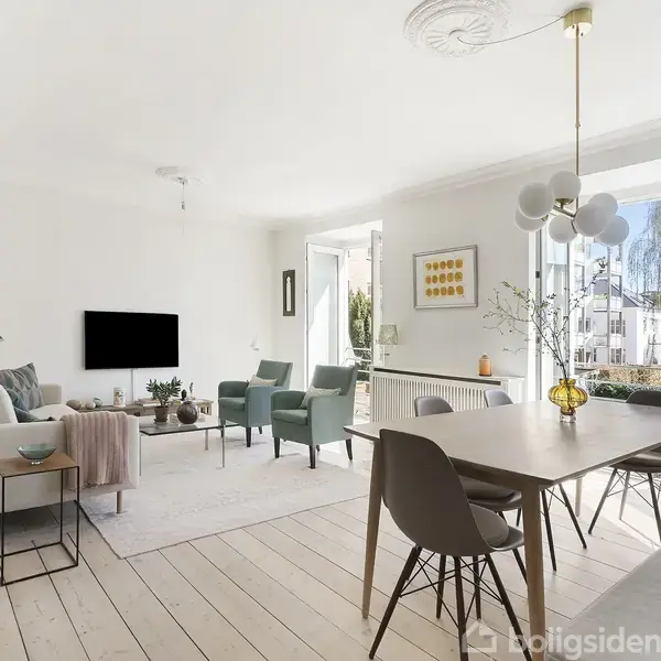 Stue indrettet med lyse træplanker, en sofagruppe med sofa og stole, spisebord med stole. Lysindfald fra åbne dobbeltdøre til balkon. Grøn plante og moderne lamper til stede.