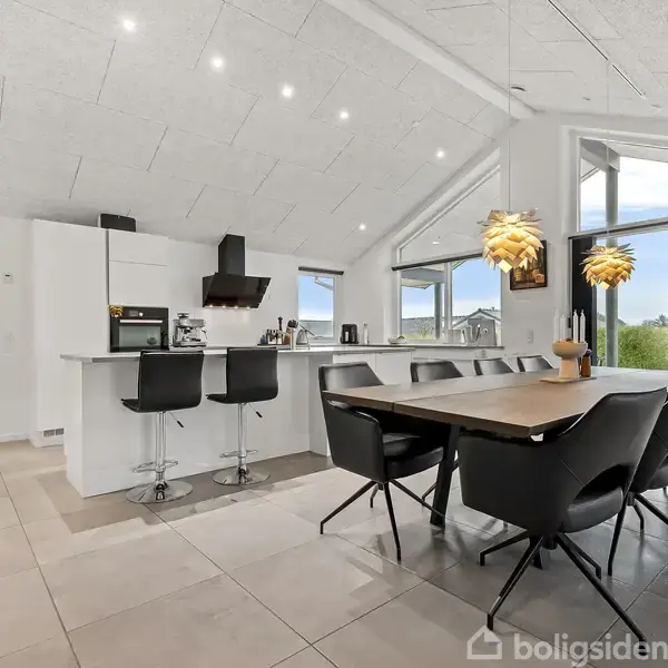 Et moderne køkken og spisestue er placeret i et rummeligt, lyst rum med hvide vægge og loft. Køkkenøen har barstole, og spisebordet er omgivet af sorte stole. Store vinduer viser en grøn udsigt udenfor.