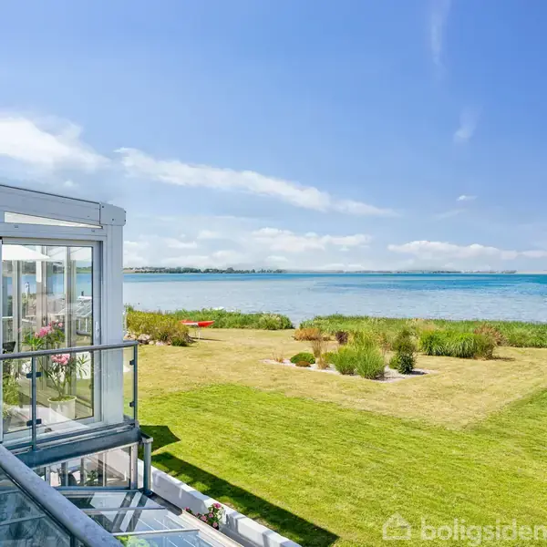 Glasveranda med udsigt står stille ved en frodig græsplæne og kystlinje ud til en rolig sø. Skyfri blå himmel giver en lys og indbydende atmosfære.