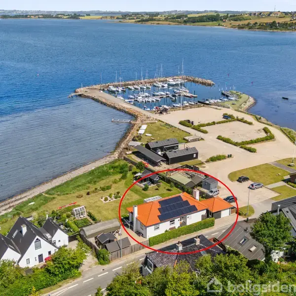 Et hus med rødt tegltag og solpaneler er omkranset med en rød cirkel. Det ligger nær en havn med både, omgivet af grønne områder og havudsigt.