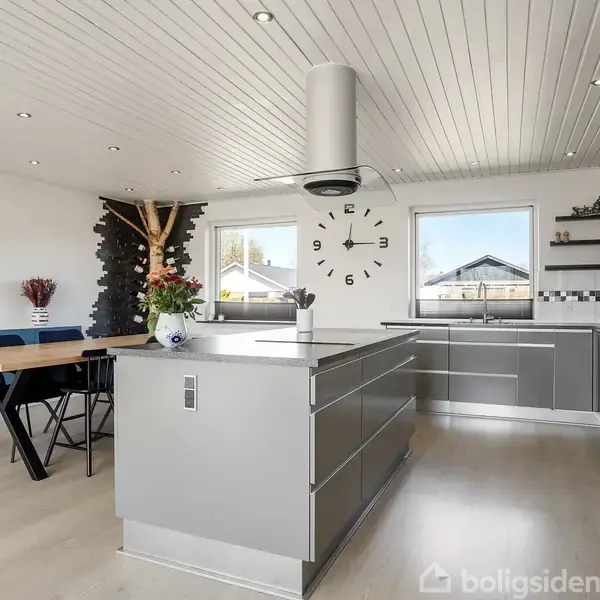 Køkkenalrum med moderne indretning. Et stort spisebord med blå stole står nær en ø-køkkenø. Vægge er hvide med et stort ur. Store vinduer lader naturligt lys ind.
